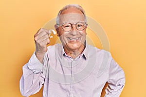 Senior man with grey hair holding medical hearing aid looking positive and happy standing and smiling with a confident smile