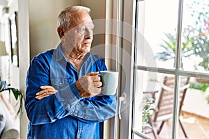 Senior man with grey hair leaning by the window of his home, drinking a cup of coffee in the morning