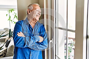 Senior man with grey hair leaning by the window of his home, smiling happy and confident looking outside