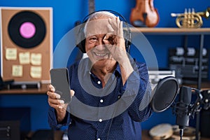 Senior man with grey hair showing smartphone screen at music studio smiling happy doing ok sign with hand on eye looking through