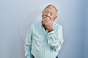 Senior man with grey hair standing over blue background bored yawning tired covering mouth with hand