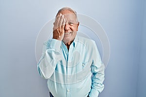 Senior man with grey hair standing over blue background covering one eye with hand, confident smile on face and surprise emotion