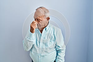 Senior man with grey hair standing over blue background tired rubbing nose and eyes feeling fatigue and headache