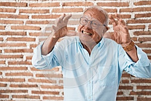 Senior man with grey hair standing over bricks wall shouting frustrated with rage, hands trying to strangle, yelling mad