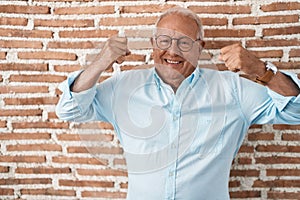 Senior man with grey hair standing over bricks wall showing arms muscles smiling proud