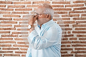 Senior man with grey hair standing over bricks wall tired rubbing nose and eyes feeling fatigue and headache