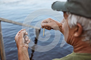 Senior man with grey hair wearing baseball cap and green t shirt baits fishing rod, elderly male spending time near river or lake