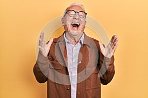 Senior man with grey hair wearing casual jacket and glasses celebrating mad and crazy for success with arms raised and closed eyes