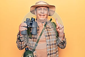 Senior man with grey hair wearing hiker bakcpack holding binoculars smiling with an idea or question pointing finger with happy