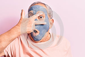 Senior man with grey hair wearing mud mask peeking in shock covering face and eyes with hand, looking through fingers afraid
