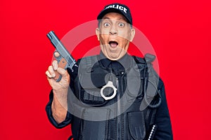 Senior man with grey hair wearing police uniform holding gun scared and amazed with open mouth for surprise, disbelief face