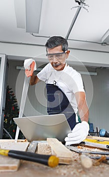 Senior man hold saw and check his project in laptop computer.