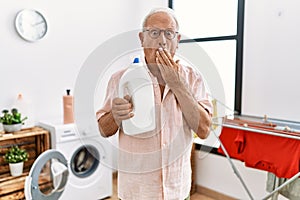 Senior man holding detergent bottle at laundry room covering mouth with hand, shocked and afraid for mistake