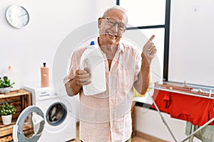 Senior man holding detergent bottle at laundry room smiling happy pointing with hand and finger to the side