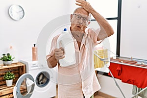 Senior man holding detergent bottle at laundry room stressed and frustrated with hand on head, surprised and angry face