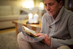 Senior man at home reading Bible, burning candles behind him