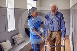 Senior Man At Home Using Walking Frame Being Helped By Female Care Worker In Uniform