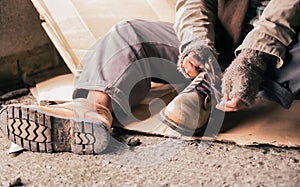 Senior man homeless wearing sweater and cloth gloves with sitting on cardboard and tying laces on shoes