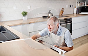 A senior man indoors in kitchen at home, loading dishwasher.