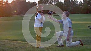 Senior man on knee giving flowers to his woman outdoors.