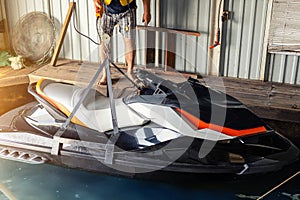 Senior man launching or lifting modern fast jetski hanged on strpas with wire hoist rail crane for dry storage. Boat garage