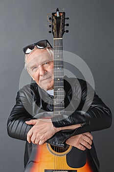 Senior man in leather jacket and sunglasses up on head standing isolated on gray hugging guitar looking camera serious