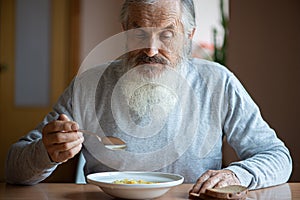 Senior  man with a long gray beard sitting by the table and eating soup and bread