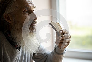 Old sad man with a long gray beard sitting by the window and eating bread