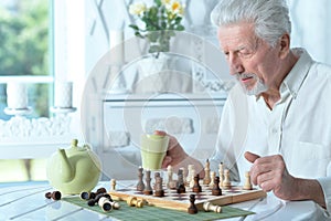 Senior man looking at chess board while drinking tea at home