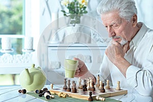 Senior man looking at chess board while drinking tea at home