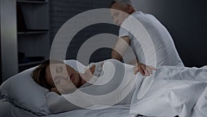 Senior man looking at sleeping wife, male sitting on far bed edge backwards