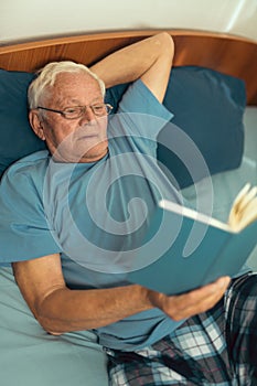 Senior man lying on bad and reading book