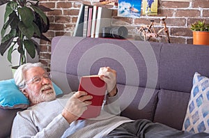 Senior man lying on sofa and reading book at home - retired, leisure and people concept