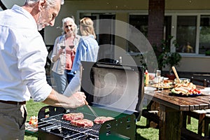 Senior man making barbecue at outdoor grill while women drinking wine behind