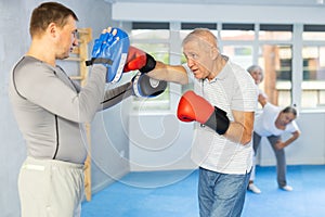 Senior man and male couch are boxing in gym, coach helps student to work out force of blow.