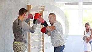 Senior man and male couch are boxing in gym, coach helps student to work out force of blow.