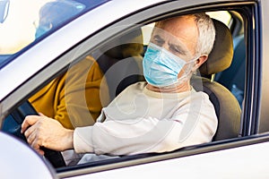 Senior man in a medical face mask driving a car. Coronavirus pandemic concept. Road trip, travel and old people concept - happy se