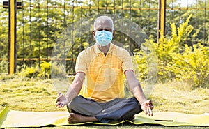 Senior man in medical face mask meditating on yoga mat at park - cconcept of elderly people healthcare, fitness during coronavirus