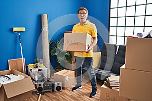 Senior man moving to a new home holding cardboard box in shock face, looking skeptical and sarcastic, surprised with open mouth