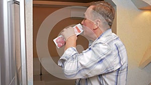 Senior man opens the fridge and drink the juice from the package. It quenches thirst and puts juice back in the fridge.