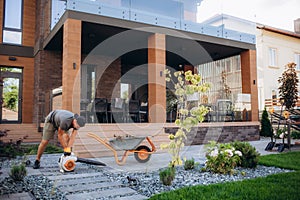 Senior man in overall working in garden with leaf blower and daughter mowing lawn in background in autumn