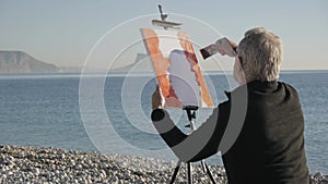 Senior man paints a picture on the beach. Back view of elderly male artist painting the canvas with a wide spatula at