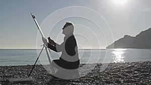 Senior man paints a picture on the beach. Elderly male artist painting the canvas on metal easel at beach against