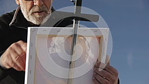 Senior man paints a picture on the beach. Low angle view of elderly male artist painting the canvas with a wide spatula