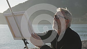 Senior man paints a picture on the beach. Mid shot of elderly male artist painting the canvas at sea beach on sunset