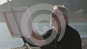 Senior man paints a picture on the beach. Mid shot of elderly male artist painting the canvas at sea beach on sunset