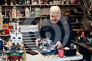 Senior man, pensioner holding made handicraft scale model of a tank on the table ih his garage with different materials