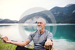 A senior man pensioner sitting by lake in nature, doing yoga exercise.