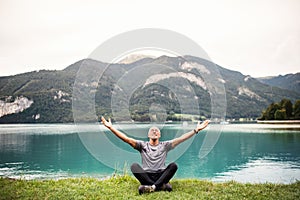 A senior man pensioner sitting by lake in nature, doing yoga exercise.