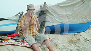 Senior man playing bamboo flute on the beach next to fishing boat